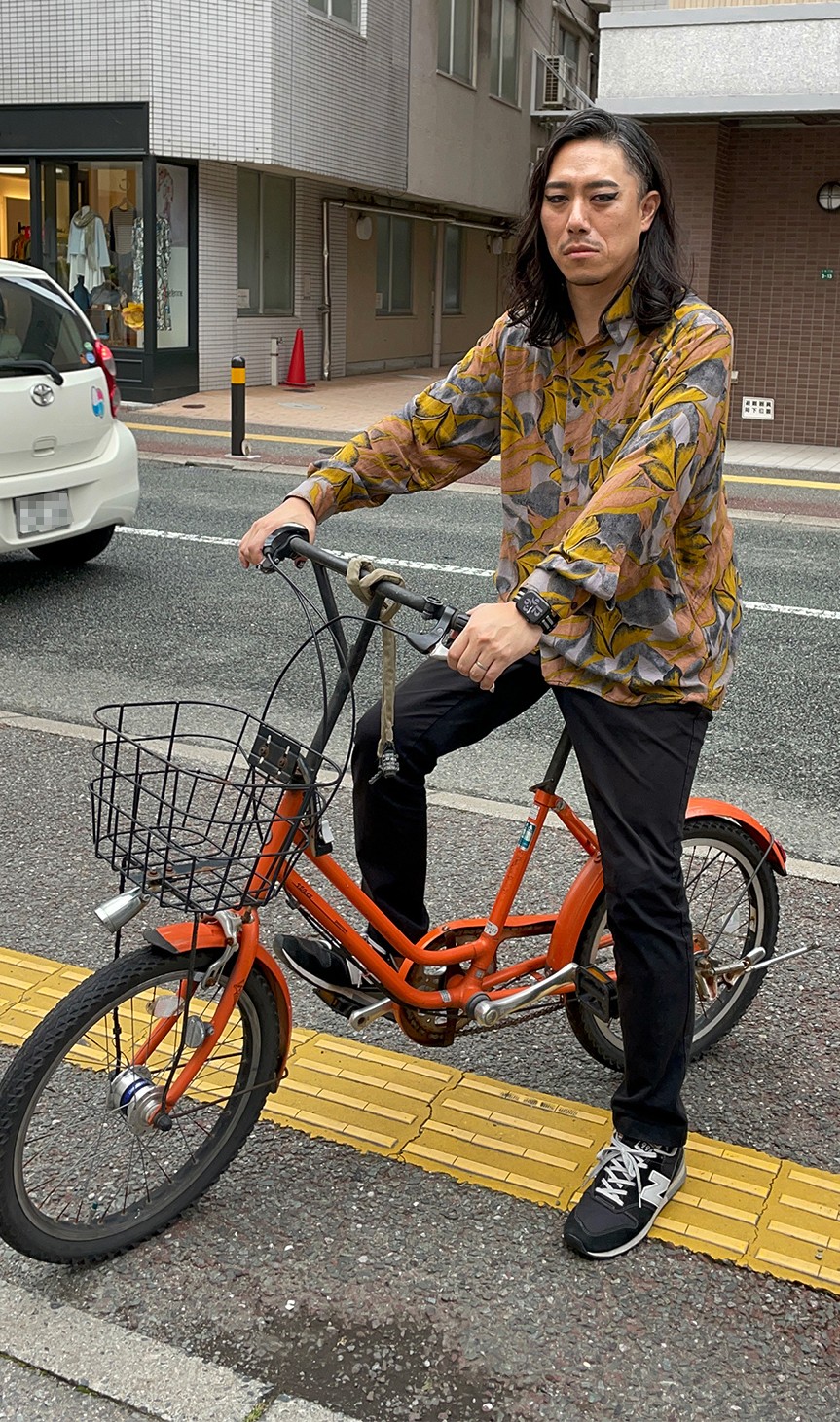「フラワーボーイ」カットの妖艶なメイクのまま街中でオレンジの自転車にまたがる今村。