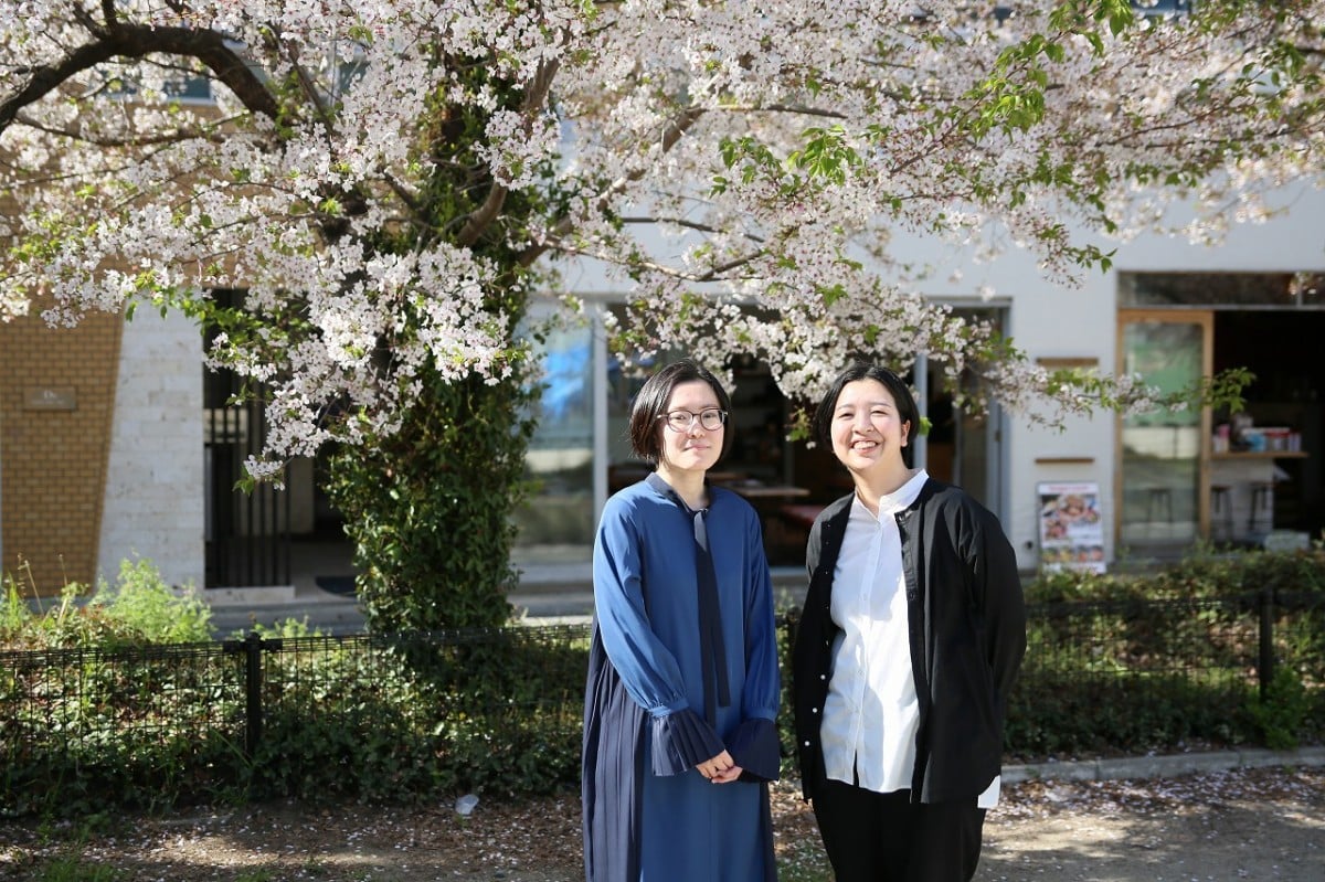 桜の木の下で新卒の小牧さんと三隅さんが笑顔でこっちを見ている様子