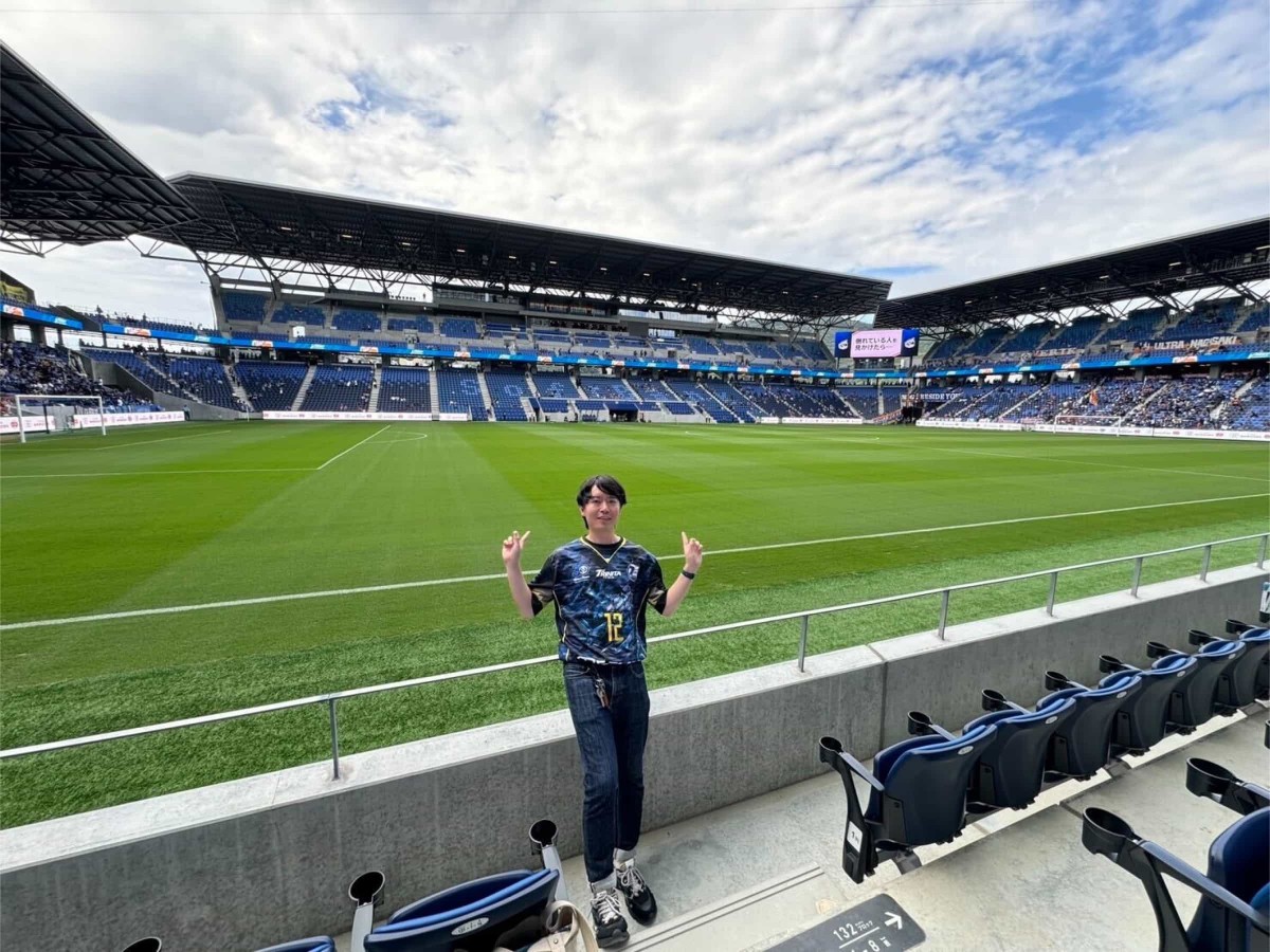 ピーススタジアムのグラウンドをバックにピースサインをする佐藤さん