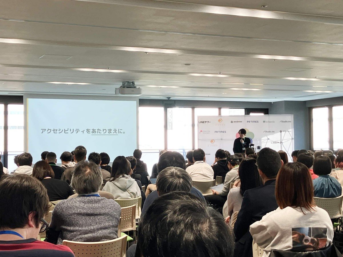ディーゼロ・吉田さんのスポンサーセッションの様子。写真手前には座ってセッションを聞くたくさんの人々。奥のスクリーンには「アクセシビリティをあたりまえに。」という文字が映し出されている。その隣の演壇で、吉田さんが話している。