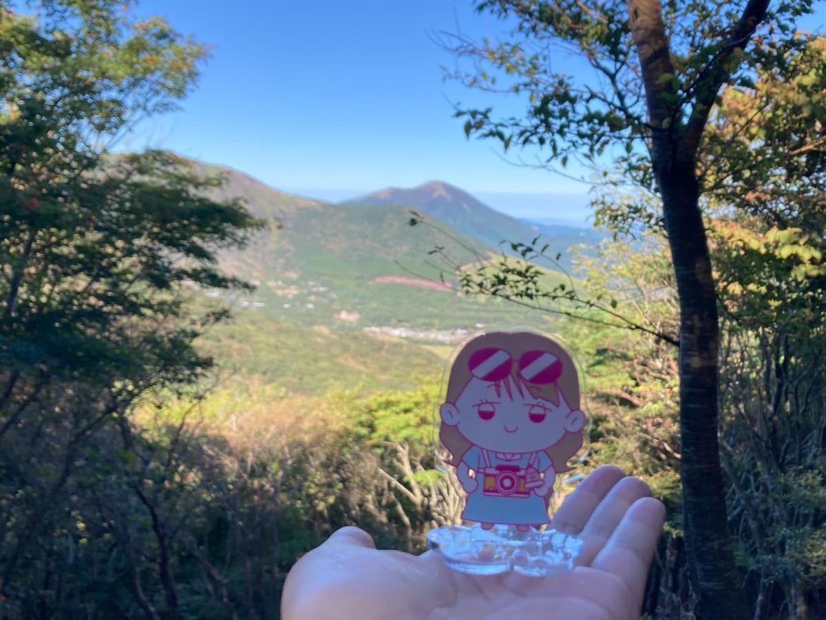 登山中の眺望と手のひらに載せられたアクリルスタンドのヨネチ。ヨネチの向こう側にくじゅうの山々の景色が広がる。