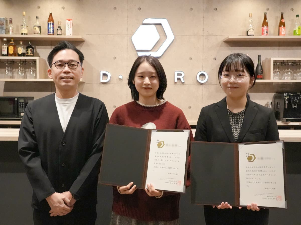 ディーゼロ社長と、社長の隣に立つ内定者2人。内定者の2人は内定証書を手で広げて持ち、3人とも微笑んでいる。