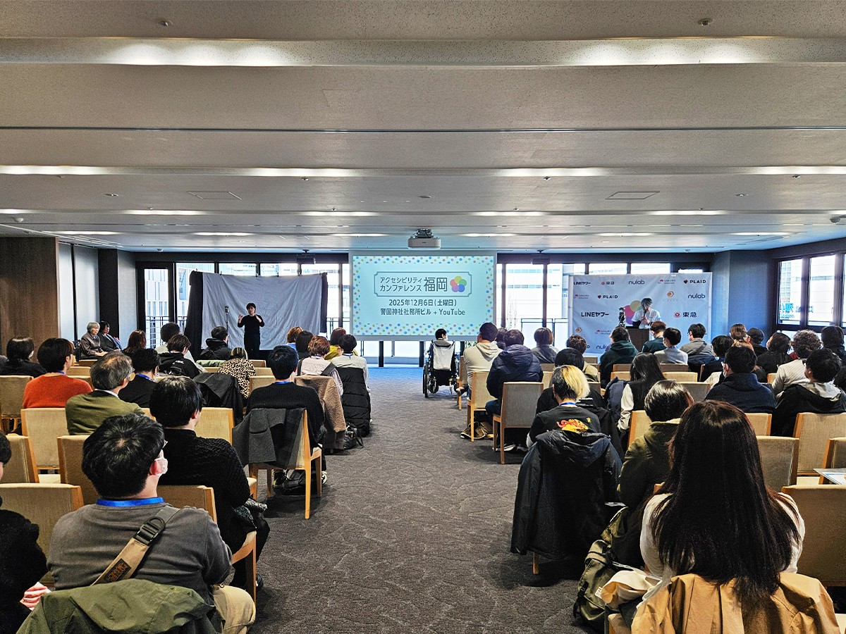 アクセシビリティカンファレンス福岡2025の会場内の様子。多くの参加者が着席して前方のスクリーンに注目している。スクリーンの左側には手話通訳者が立ち、右側には司会者のブースがある。
