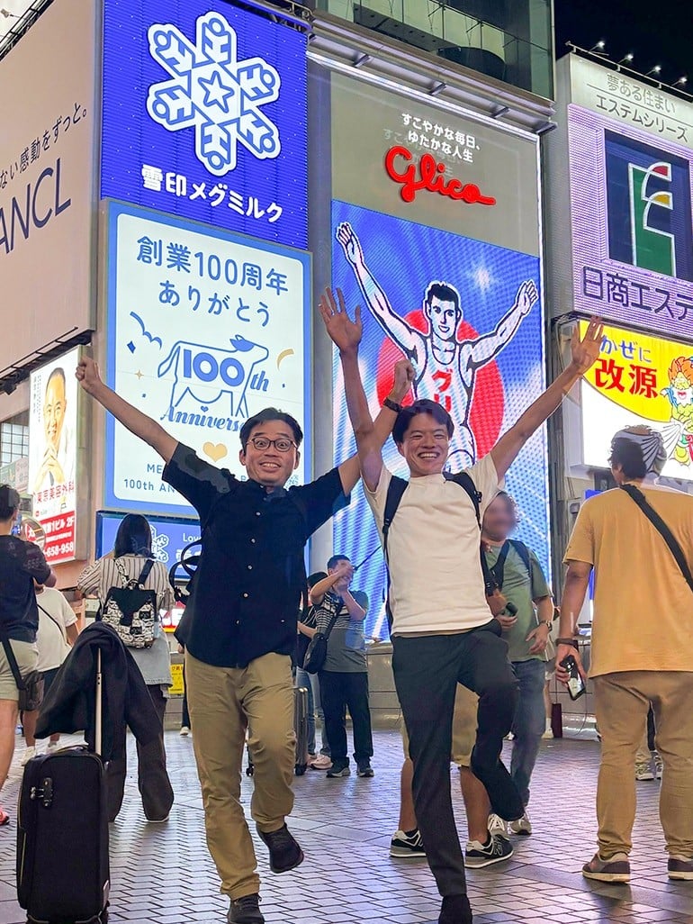道頓堀グリコサインの前で、谷口さんと小林さんが並んでグリコのポーズを取っている。