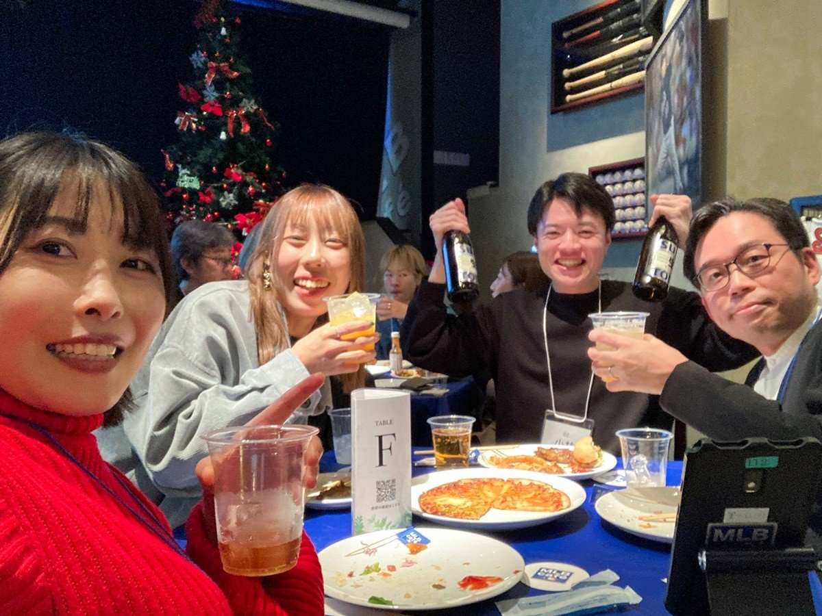 忘年会のテーブルでの写真。料理がならんだテーブルを囲んだメンバーがボトルを両手を手に持ったり、お酒の入ったカップを手に楽しそうにしている。