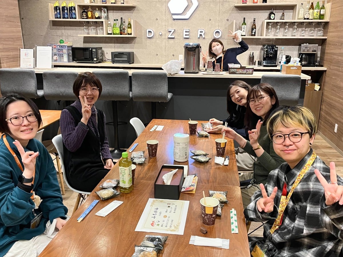 オフィスのカフェエリアで、おにぎりや飲み物が置かれたテーブルを囲んだスタッフと、その後ろで飲み物を準備するスタッフがカメラに向けてピースしている。