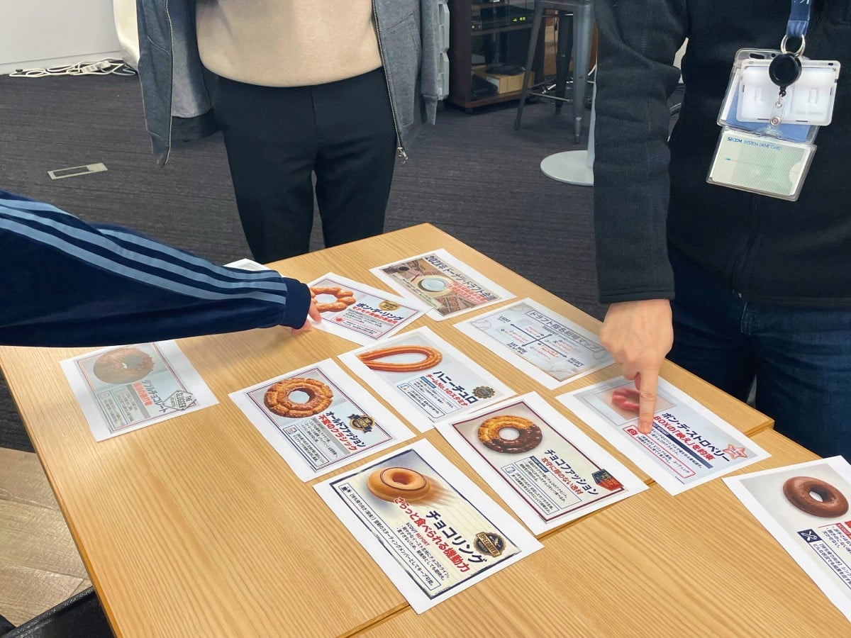 各ドーナツの特徴が記載された紙がテーブルの上に並べられており、スタッフ2名が食べたいドーナツの紙に指を指している。