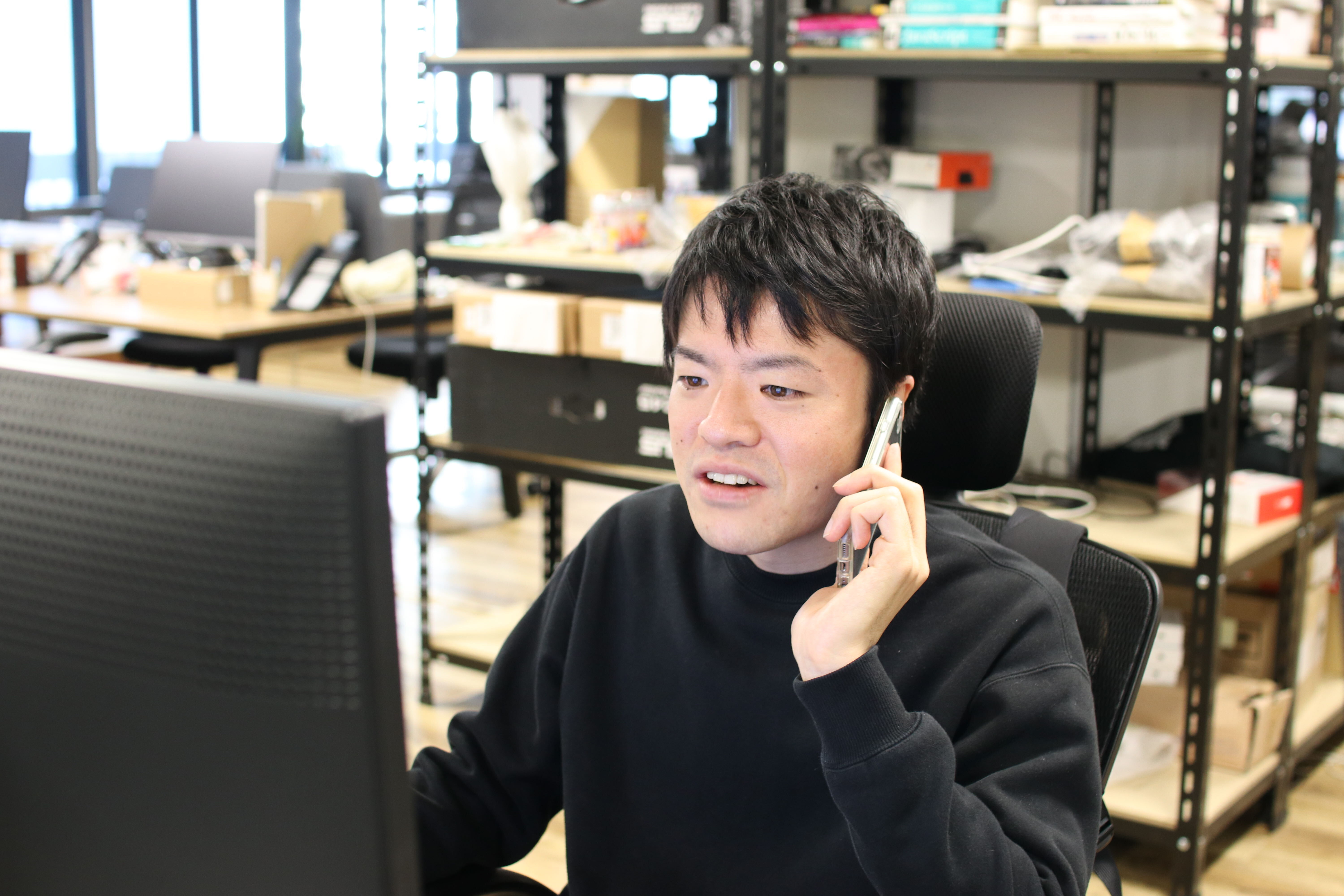 オフィスで小林さんが電話対応を行っている。PCを見ながら、右手にはスマートフォン持っている。