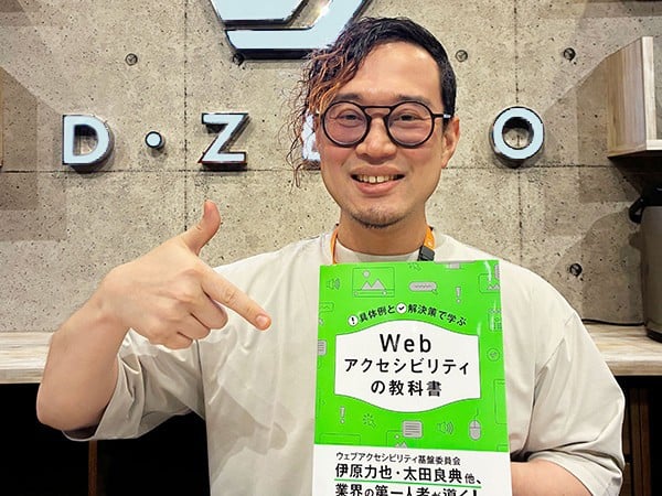 『具体例と解決策で学ぶ Webアクセシビリティの教科書』を手に持ち、笑顔で指差す平尾
