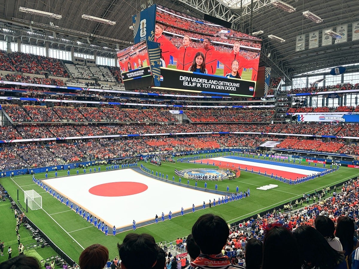 満員のサッカースタジアムで行われている日本対オランダの試合前セレモニー。ピッチに広げられた両国の巨大な国旗。