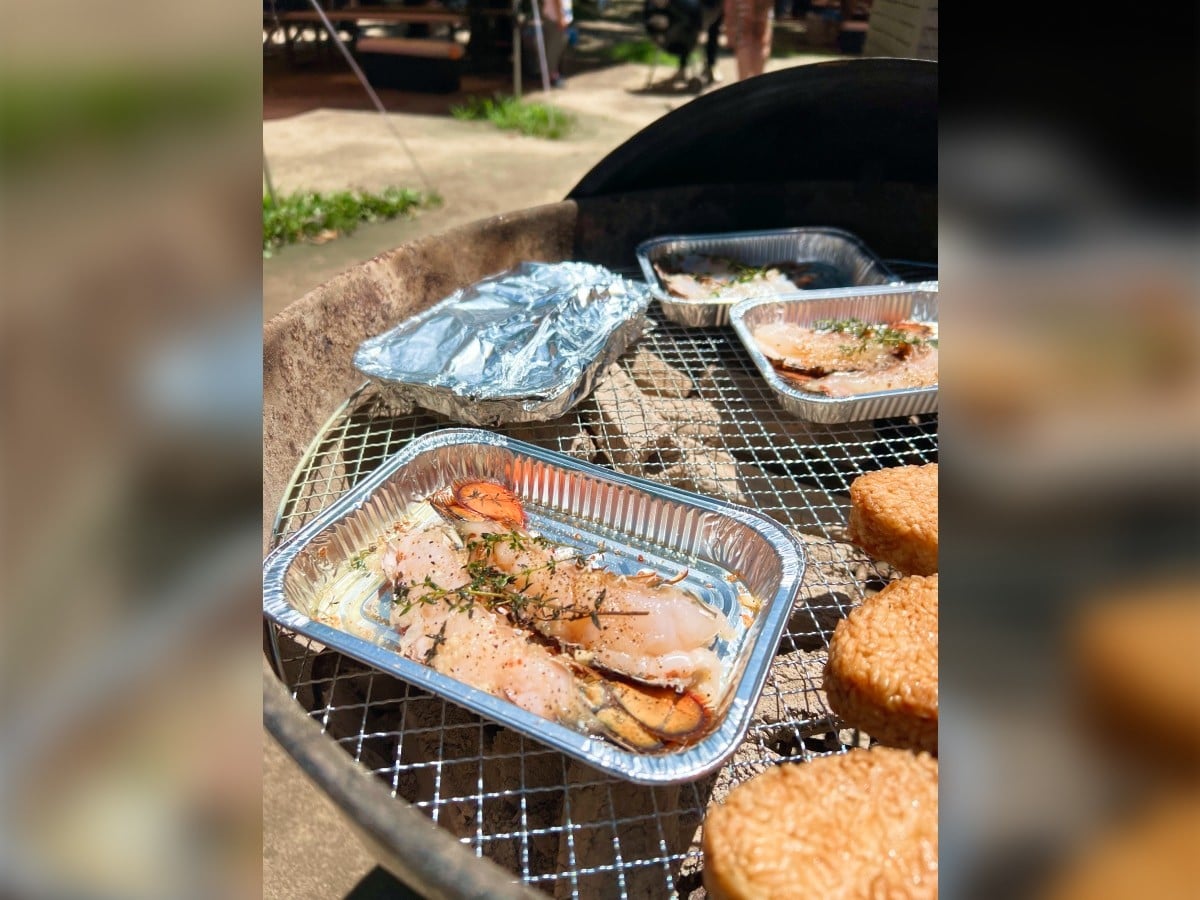 BBQグリルで調理中のハーブを添えた魚介類のアルミトレーと、その隣に並ぶ焼きおにぎり。