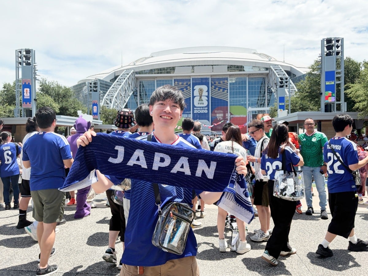 スタジアムの前でこちらを見て笑っている佐藤さん。両手で広げているタオルには「JAPAN」の文字が書かれている。