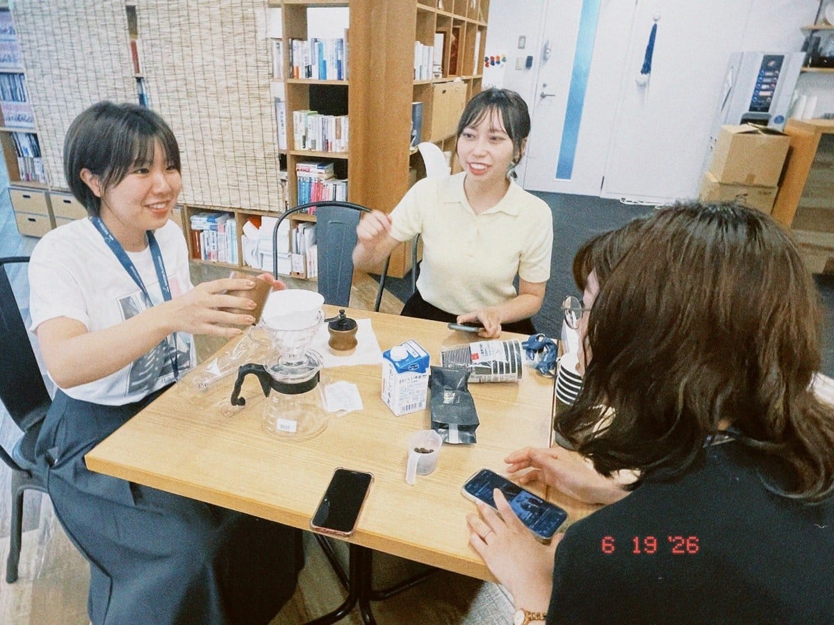 オフィスの休憩スペースで、木製のテーブルを囲んで3人の女性社員が笑顔で談笑している様子。テーブルの上にはハンドドリップコーヒーの器具や牛乳パック、スマートフォンが置かれている。