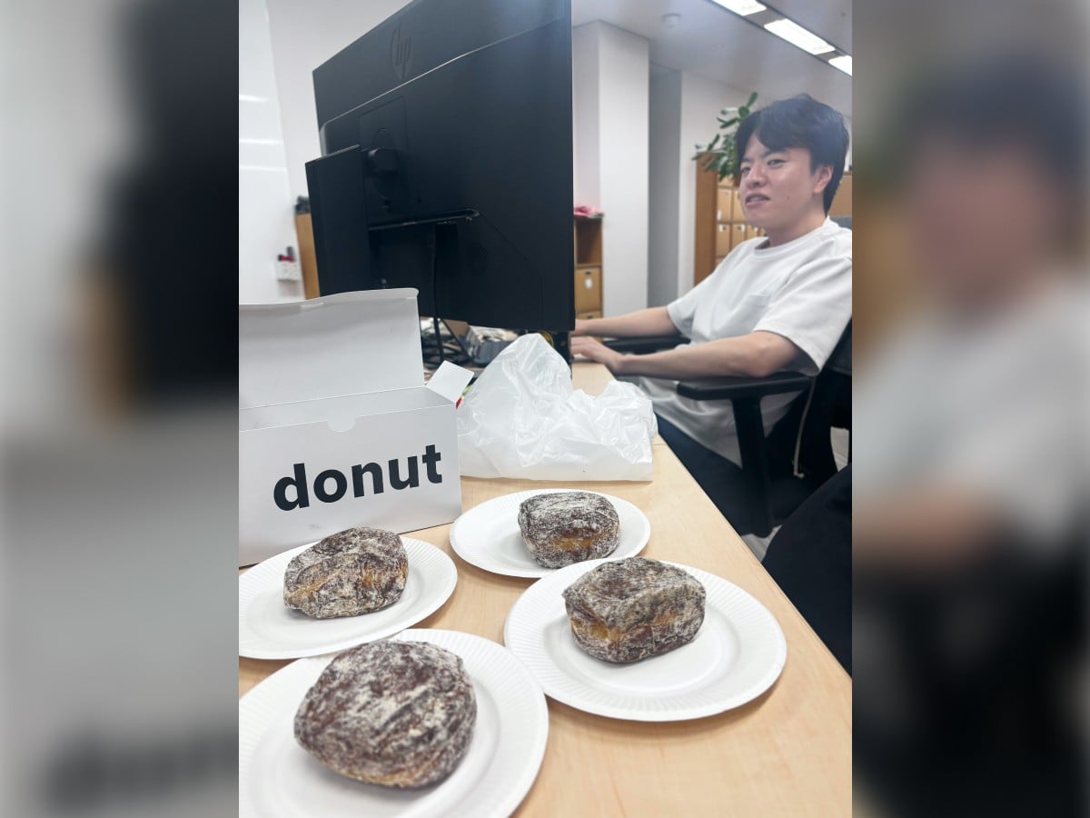 オフィスのデスクの上に、紙皿に載せられた4つのシュガードーナツと白い箱が置かれている様子。デスクの奥には大きなPCモニターがあり、白いTシャツを着た小林さんが椅子に座って作業をしながら、こちらに視線を向けている。