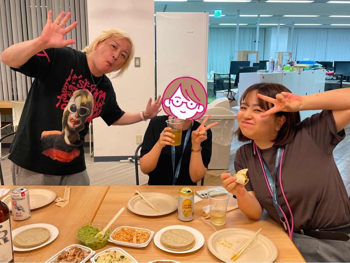 オフィスの一角で、テーブルに食事や飲み物を広げて楽しそうにポーズをとる東京メンバー3人の様子。左側の男性は、金髪で、個性的な顔のイラストがプリントされた黒いTシャツを着ており、カメラに向かって身を乗り出しながら右手を大きく上げてポーズをとっています。    中央の女性は、顔がメガネをかけた女性のイラストスタンプで隠されていますが、片手に黄色い飲み物の入った透明なカップを持ち、もう一方の手でピースサインをして座っています。    右側の女性は、茶色いボブヘアでブラウンのTシャツを着ており、首からピンク色のネックストラップを下げています。片手にタコスを持ちながら、顔の横でピースサインを作って笑顔でカメラを見ています。    手前の木製のテーブルには、お酒の缶、トルティーヤの皮、ワカモレ（アボカドのディップ）、おかずが乗った紙皿などが並べられています。背景にはPCモニターが並ぶデスク群が見えます。オフィス内でカジュアルなタコスパーティーが行われており、アットホームな雰囲気が伝わってきます。