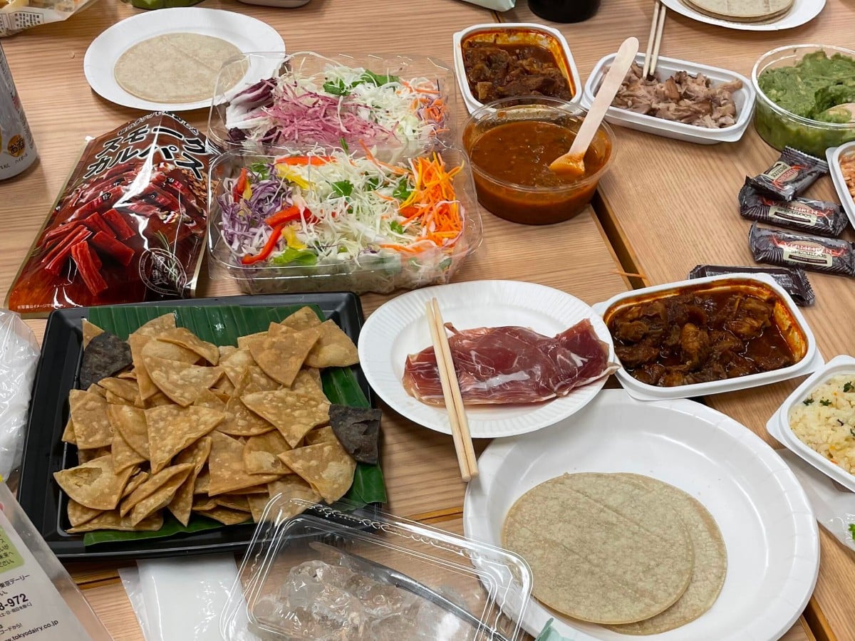木のテーブルの上に所狭しと並べられた、タコスパーティー用の様々な食事の様子。    左側には、緑色の葉が敷かれた黒いトレーにたっぷりと盛られたトルティーヤチップスや、「スモークカルパス」と書かれた赤いおつまみの大袋が置かれています。    中央には、千切りキャベツやニンジン、パプリカ、紫キャベツなどが入った色鮮やかなサラダの透明パックが2つ並び、その横には木のスプーンが添えられた赤いサルサソースの容器があります。また、中央の白い紙皿には生ハムと割り箸が乗っています。    右側・手前は、茶色いお肉の煮込みが入った長方形のパックが2つ、緑色のワカモレ（アボカドディップ）、個別包装されたチョコレートのお菓子が並んでいます。手前や奥の白い紙皿には、タコスを包むための丸いトルティーヤの皮が準備されており、氷の入った透明なパックも見えます。