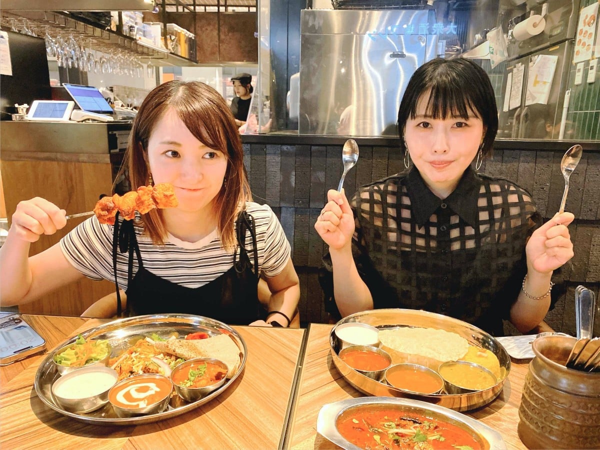 明るいカレー店内で、最近福岡にオープンした話題のカレーを前にして、楽しそうにポーズをとる「ゆるカレー部」の2人のメンバー（右から田中さん、おのさん）のツーショット写真。田中さん（前髪を切り揃えた黒髪ボブヘアで、黒いシアー素材の格子柄シャツを着た女性）は、両手にそれぞれ小さなスプーンを掲げ、笑顔でカメラを見ています。 おのさん（茶髪のボブヘアで、白黒のボーダーTシャツに黒いキャミソールを重ね着した女性）は、手元の串に刺さったチキンのようなおかずをフォークで持ち上げています。 2人の前のテーブルには、それぞれ銀色の丸いプレート（大皿）に盛られたカレーが置かれています。 田中さんの大皿（右）には、時計回りに、ナン、4つの小さな器に入った異なる種類のカレー、ヨーグルトが並んでいます。手前には赤みのあるカレーが入った金属製の器もあります。 おのさんの大皿（左）には、ナン、3つの小さな器に入った異なる種類のカレー、サラダが並んでいます。 背景には、厨房と客席を仕切る黒いタイル調の壁や、黒いTシャツを着たスタッフ1名の姿、レジ周辺の機器などが写り込んでいます。