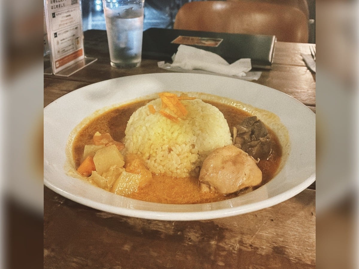 木目調のテーブルの上に置かれた、白いお皿に盛られたスパイスカレーのクローズアップ写真です。    中央にはライスがドーム状に盛られ、周りには、スープ状のサラサラとしたカレーソースがたっぷりと注がれています。    ライスの手前右側には大きくカットされた柔らかそうなチキンがゴロッと添えられ、左側にはジャガイモやニンジン、右奥には濃い茶色に煮込まれた具材が配置されています。    カレーの奥には、氷の入ったお水のグラス、黒いメニューボード、アクリルスタンドに入った卓上メニューなどが置かれています。