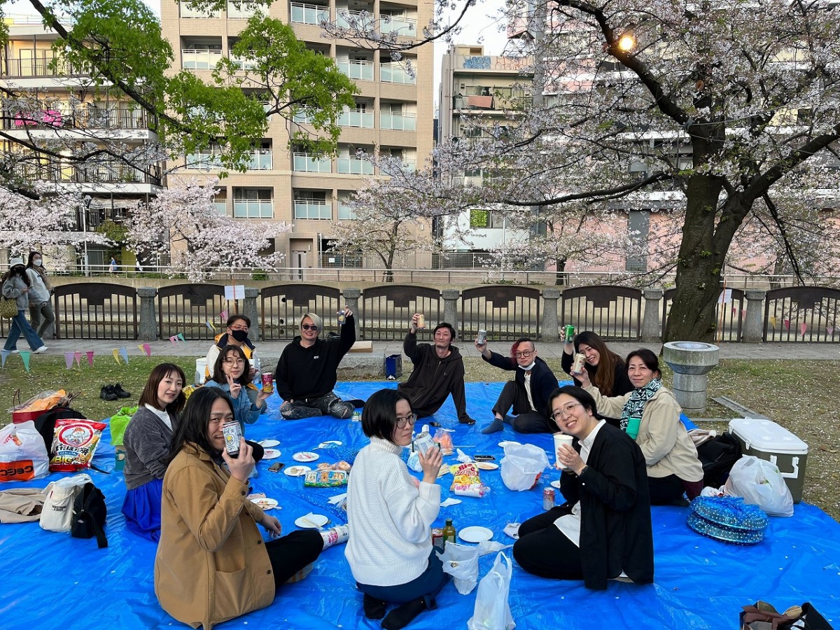 桜の下でスタッフが円になり、飲み物を持って笑っている写真