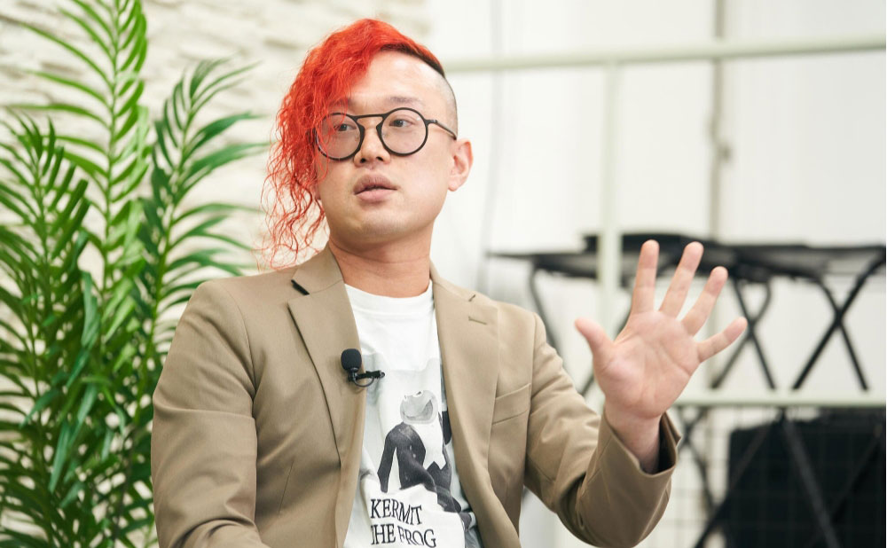 平尾の写真。髪を赤に染めたロックなヘアスタイルと、Tシャツにジャケットを着て、何かを説明している様子。